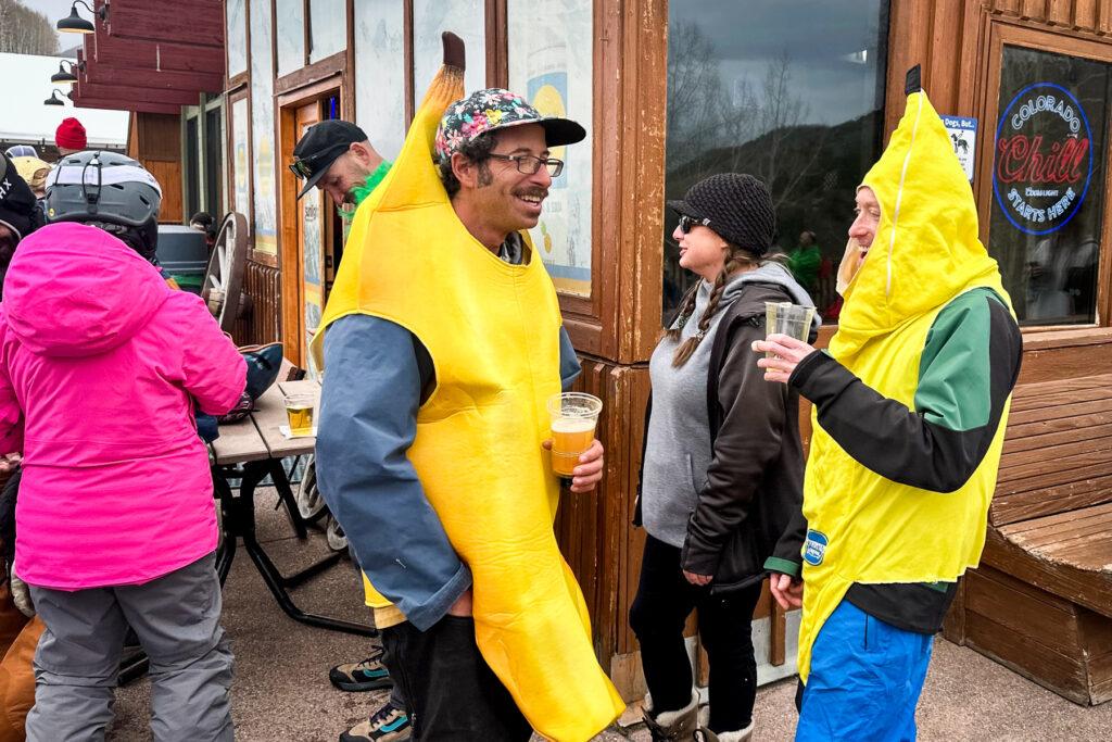 Двое мужчин одевались как бананы смеются и пиют пиво на палубе на Sunlight Ski Resort.