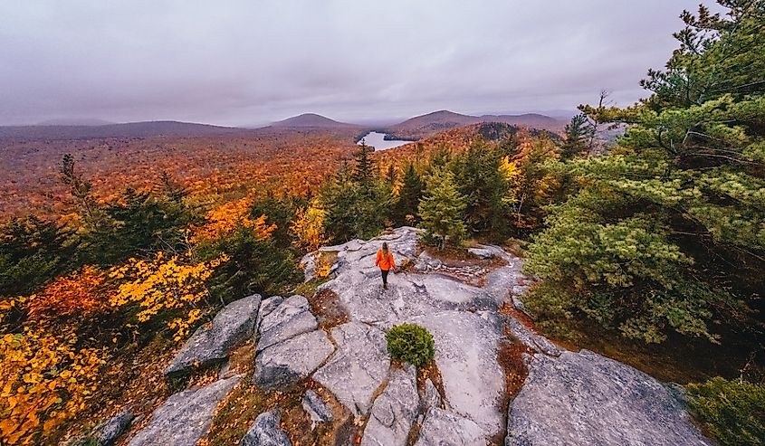 Эпический широкий пейзаж Новой Англии с осенней листвой. Воздушная женщина-путешественница в оранжевом пальто стоит на голове совы. Гора Вермонт с видом на красочные деревья осени.