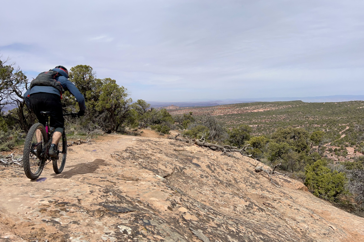 Горный велосипедист едет по каменистой тропе на фоне обширного живописного пейзажа, зеленого кустарника и далеких холмов под облачным небом.