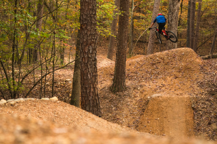 Горный байкер в синей рубашке и красном шлеме осенью совершает прыжок по грунтовой рампе в лесной местности, на деревьях видны разноцветные листья.