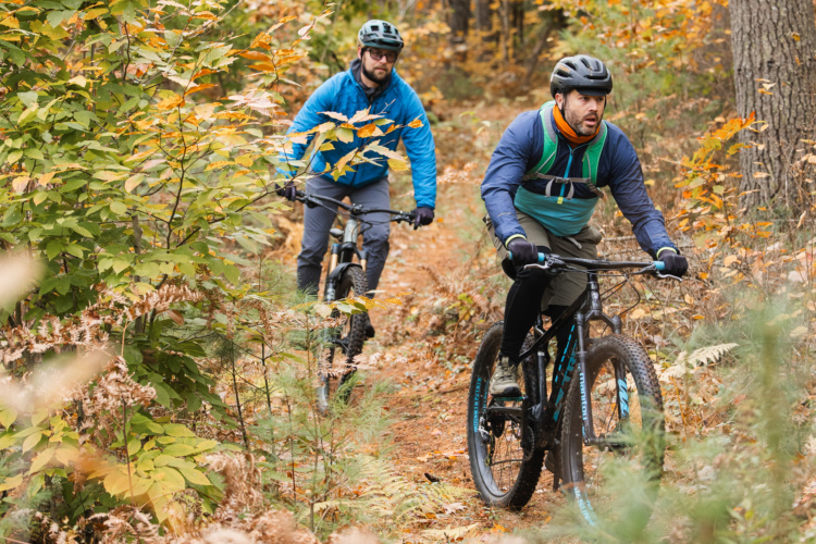 Два горных велосипедиста едут по тропе, окруженной осенней листвой. Гонщики одеты в шлемы и уличное снаряжение: один в синей куртке, другой в черно-зеленой одежде. Тропа усыпана яркими оранжевыми и желтыми листьями, создавая красочную осеннюю сцену.