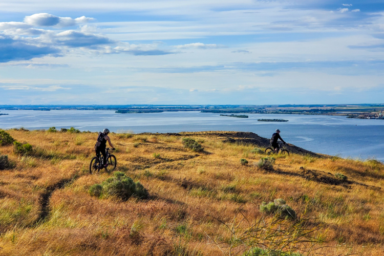 Два горных велосипедиста едут по травянистому склону холма с видом на большой водоем на фоне голубого неба и рассеянных облаков. Ландшафт включает пологие склоны и участки кустарников, подчеркивающие атмосферу приключений на свежем воздухе.
