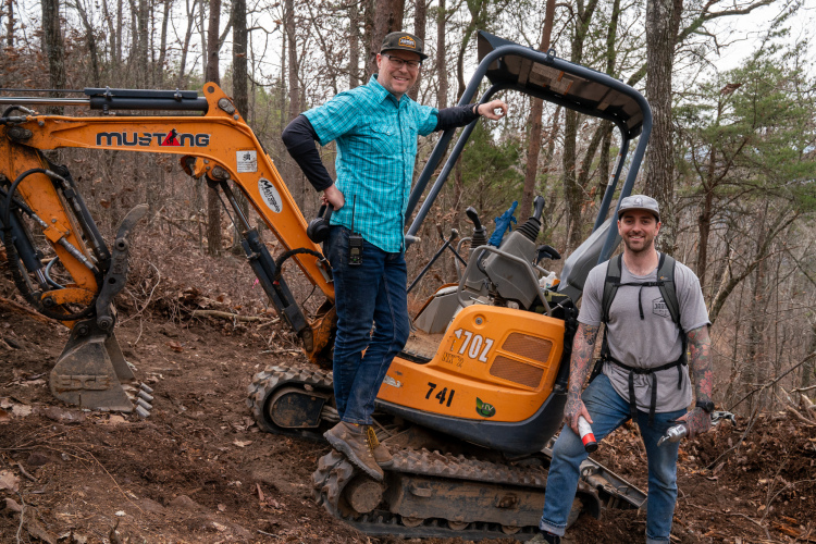 Двое мужчин позируют рядом с желто-серым мини-экскаватором в лесу. Один мужчина в синей клетчатой ​​рубашке и кепке уверенно стоит на пути экскаватора, а другой мужчина, одетый в серую рубашку с татуировками и с рюкзаком, улыбается в камеру, держа в руке инструмент. На заднем плане изображены голые деревья и разбросанные по земле листья.