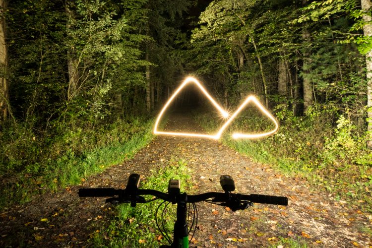 Вид с руля горного велосипеда на грунтовую тропу в лесу ночью, со светящимися световыми тропами, образующими вдалеке абстрактные горные формы. Тропа окружена деревьями и листвой, что создает спокойную атмосферу на открытом воздухе.