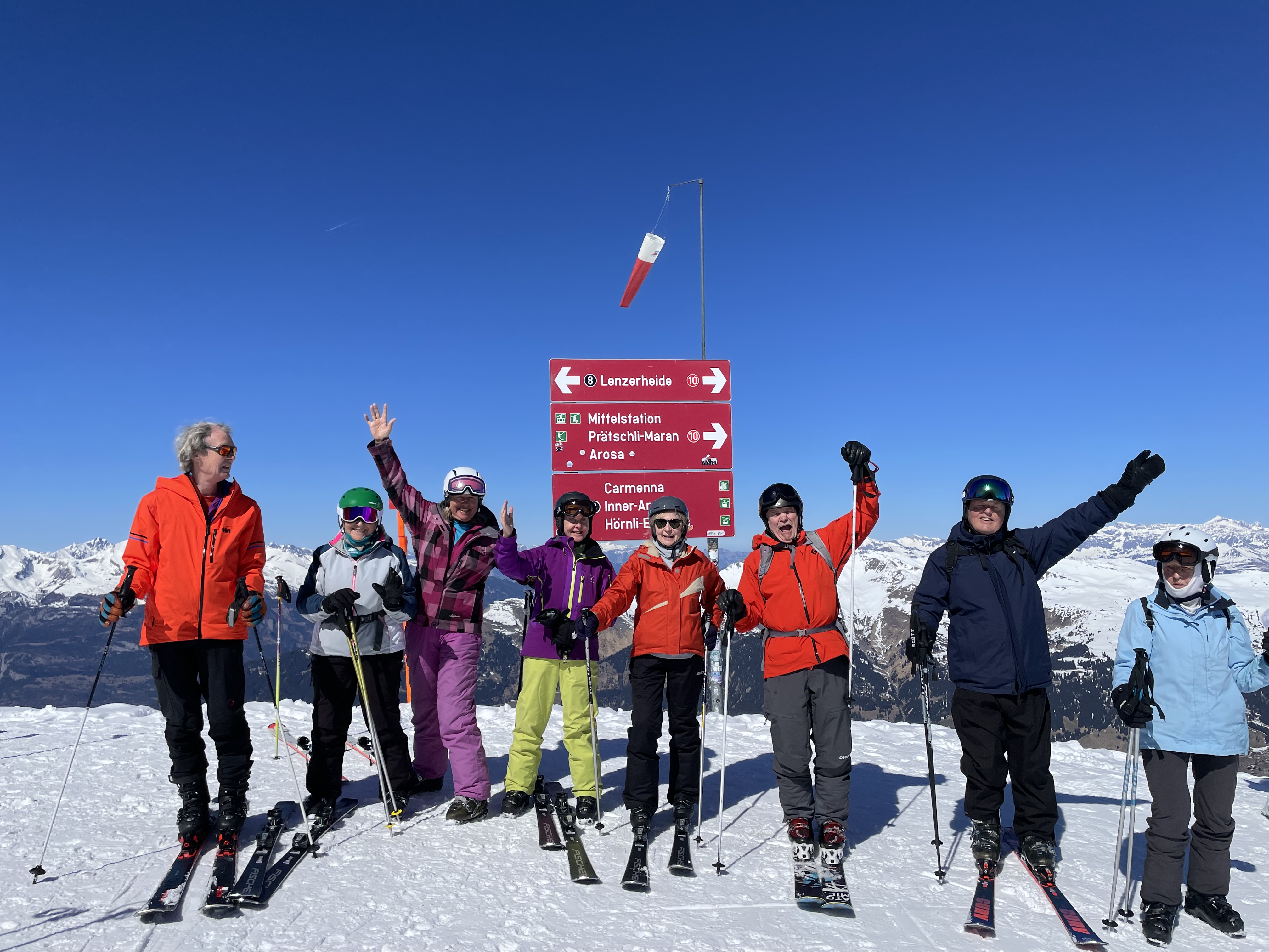 Восемь лыжников, вероятно, группа, стоят на заснеженной горе с ясным голубым небом, рядом со знаком, указывающим на Ленцерхайде и другие места.