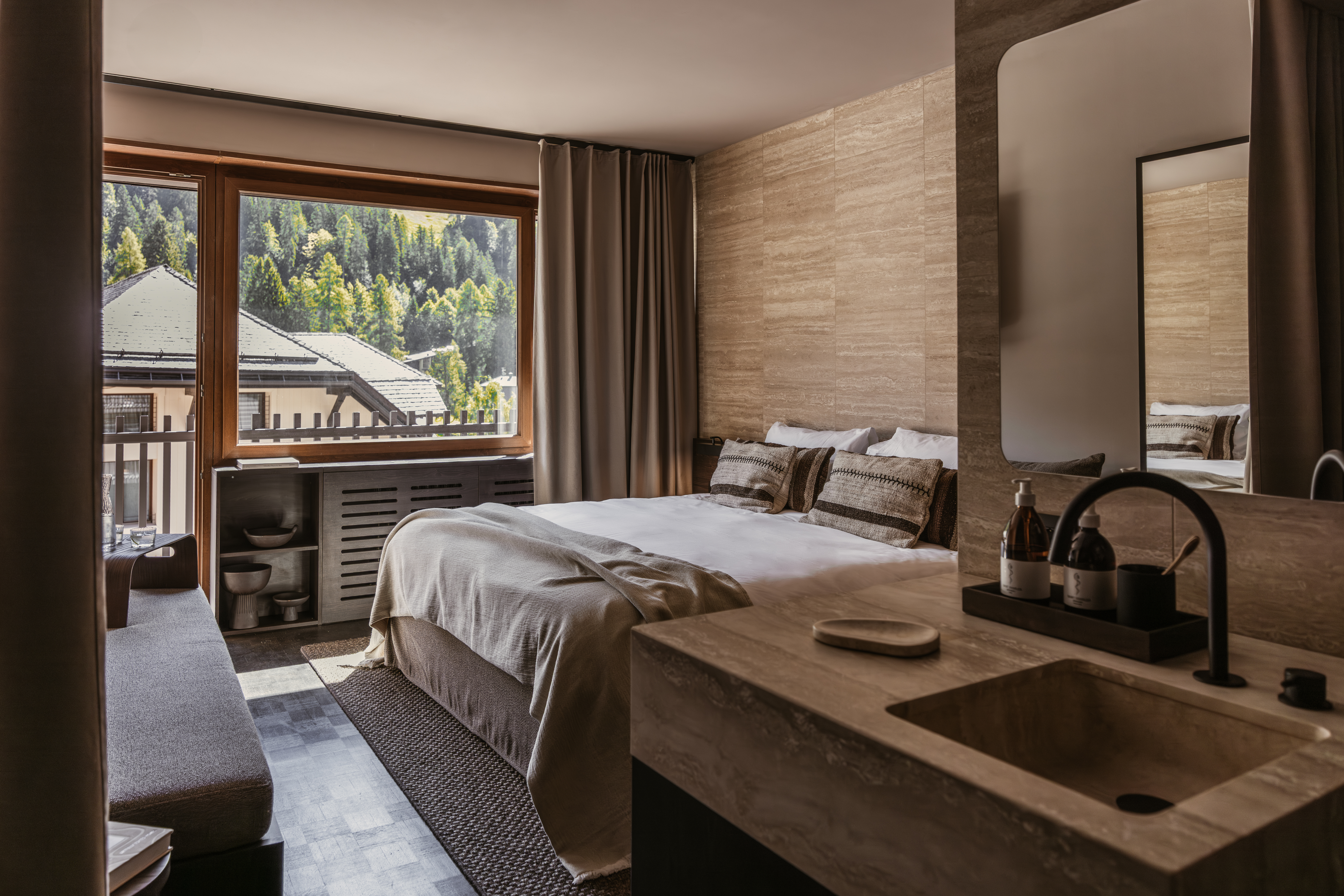 Спальня с большим окном с видом на горный лес, кроватью со светлым одеялом и подушками, а также туалетным столиком с раковиной.